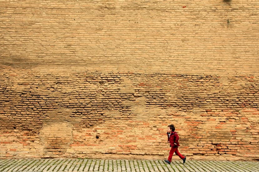 Child in a red jumpsuit running alongside a tall, weathered brick wall with visible cracks and rough textures.