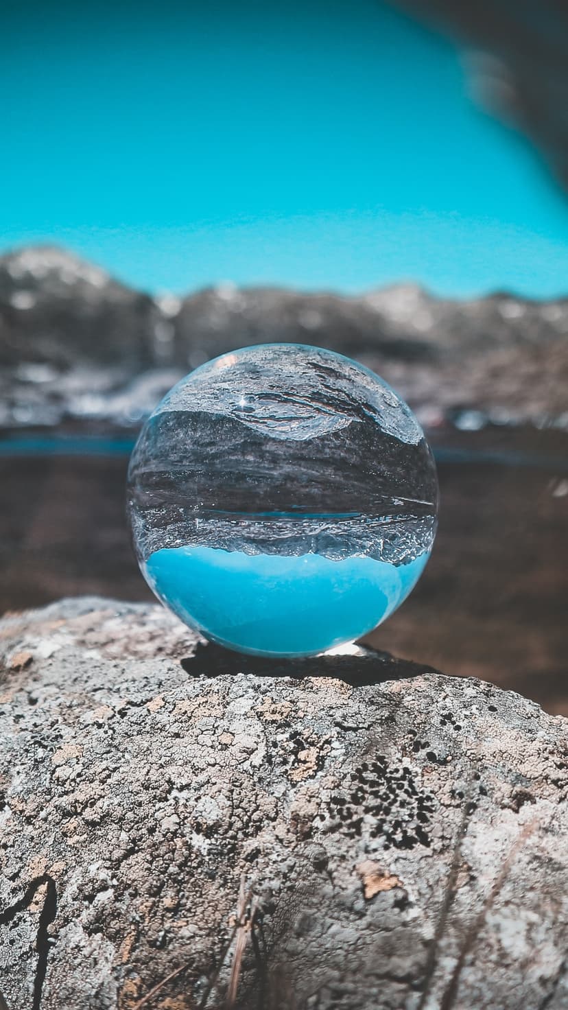 Crystal sphere on a rock captures an inverted reflection of blue mountains and a turquoise lake under a clear sky.