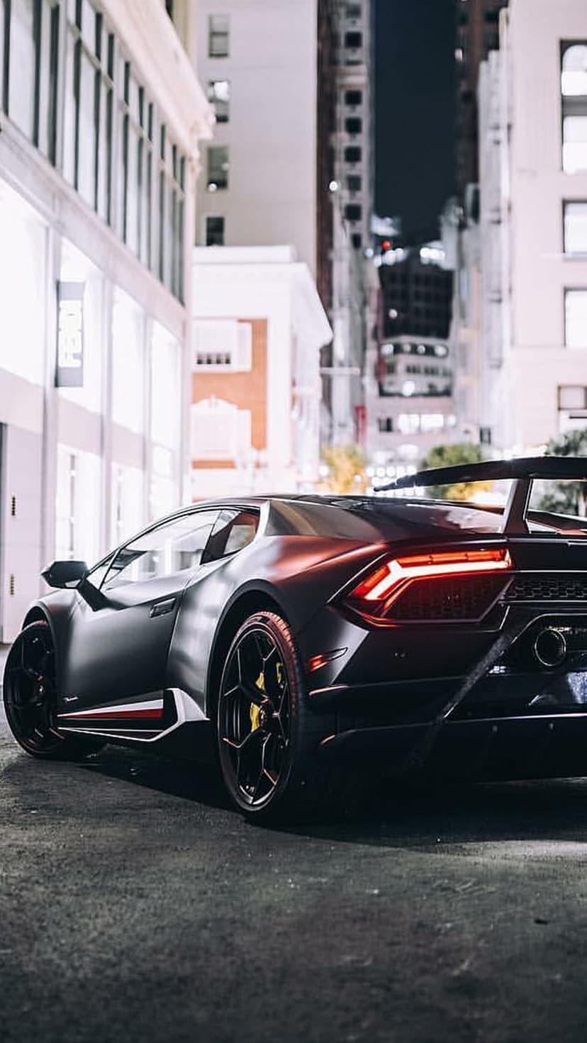 Matte black Lamborghini with red taillights parked on a dark city street with illuminated building reflections.