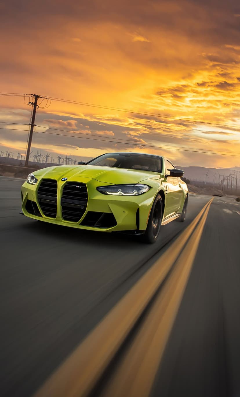 Lime green BMW M4 coupe driving on a desert road with wind turbines and a vibrant orange sunset sky.