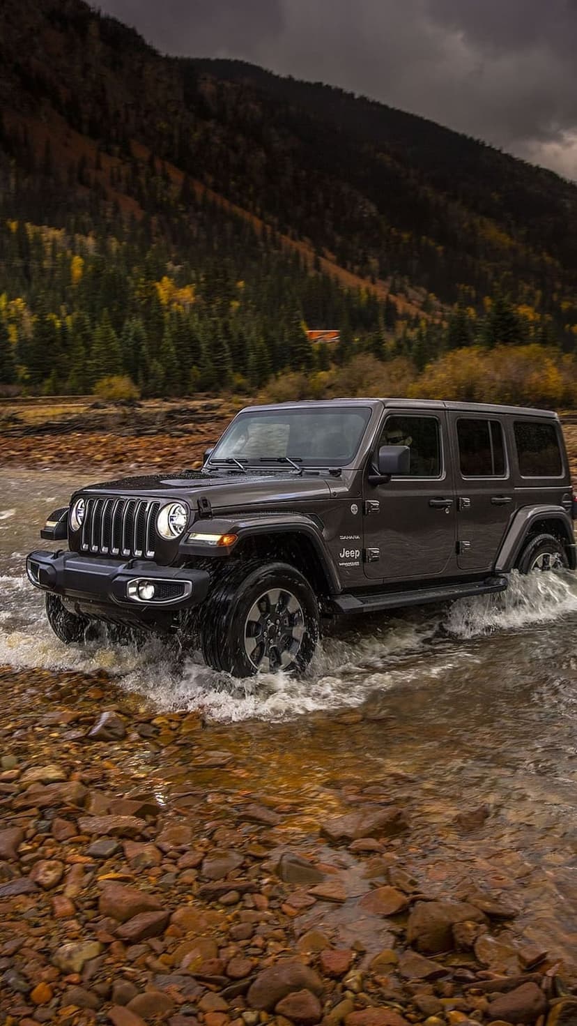 Dark gray Jeep Wrangler splashes through a shallow rocky river with autumn trees and misty mountains behind it.