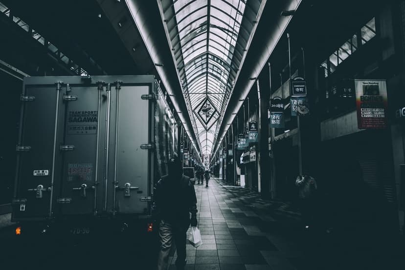 Lone man walks through a dark arcade with a geometric glass roof and dramatic noir-style urban shadows.