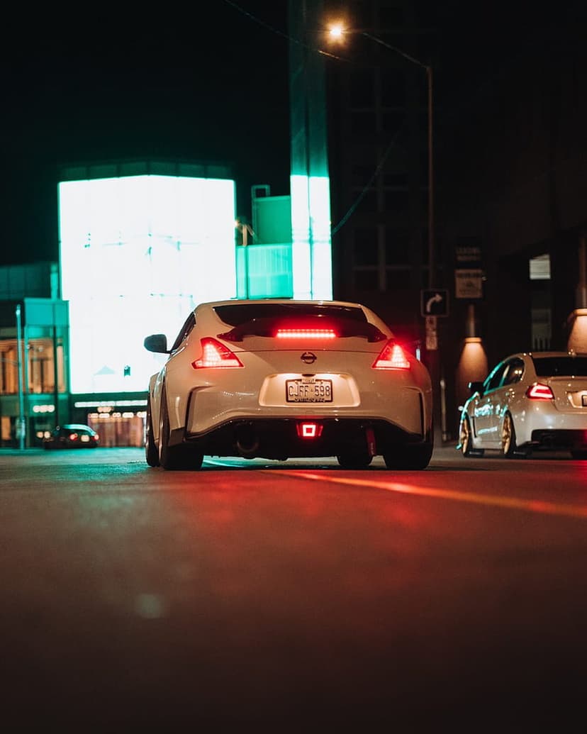 Nissan 370Z sports car traveling through a dark city street illuminated by vibrant neon signs and red lights.