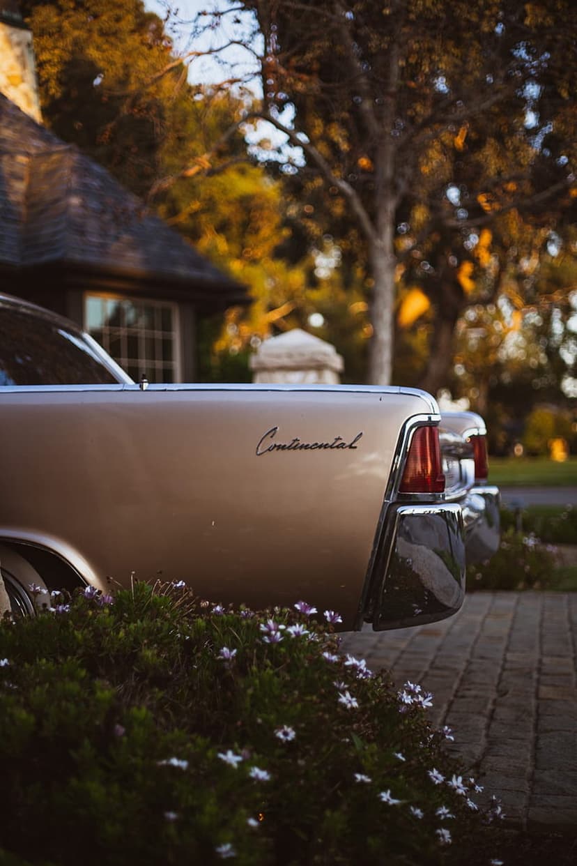 Vintage Lincoln Continental rear chrome bumper and taillights parked by a cottage garden at sunset.