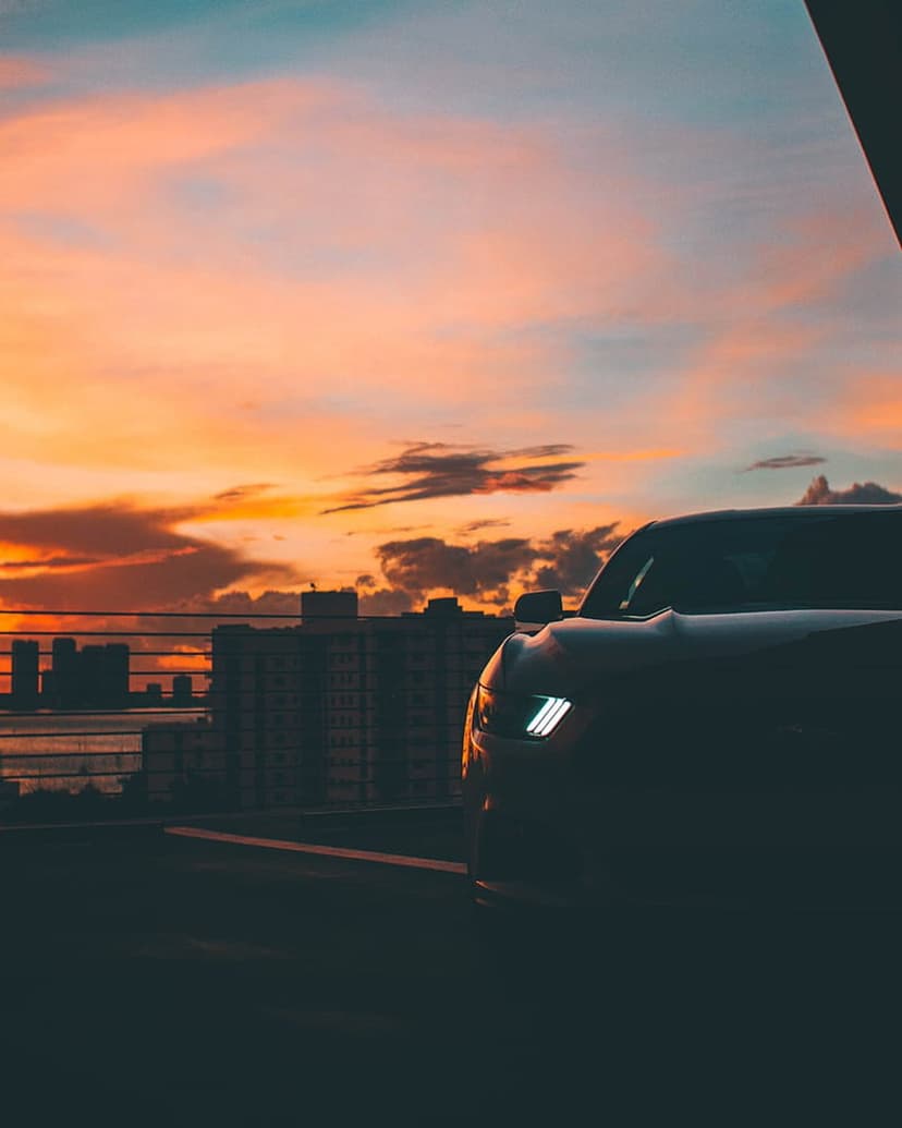 Modern car with glowing headlights sits before a city skyline under a vivid orange and purple sunset sky.