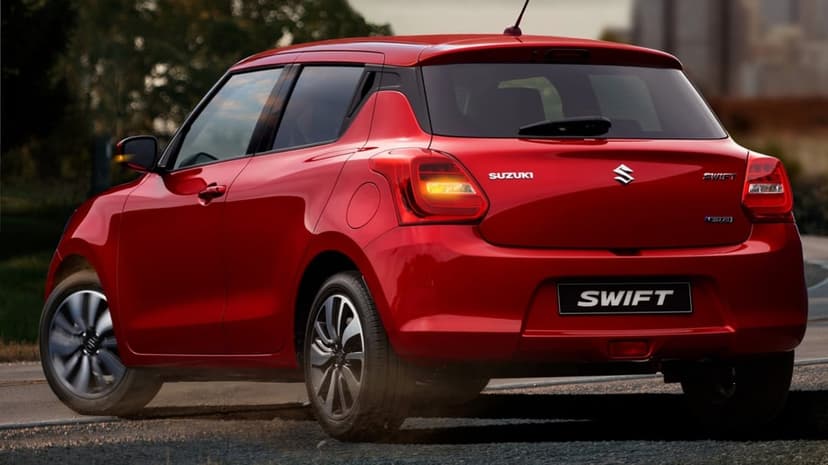 Red Suzuki Swift hatchback driving on a dusty road with wheels in motion under warm natural light.