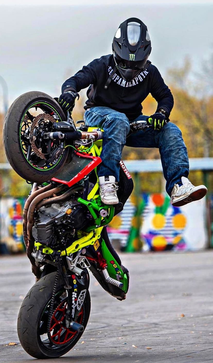 Motorcyclist in black and green gear lifts a street bike into a wheelie against a blurred mural background.