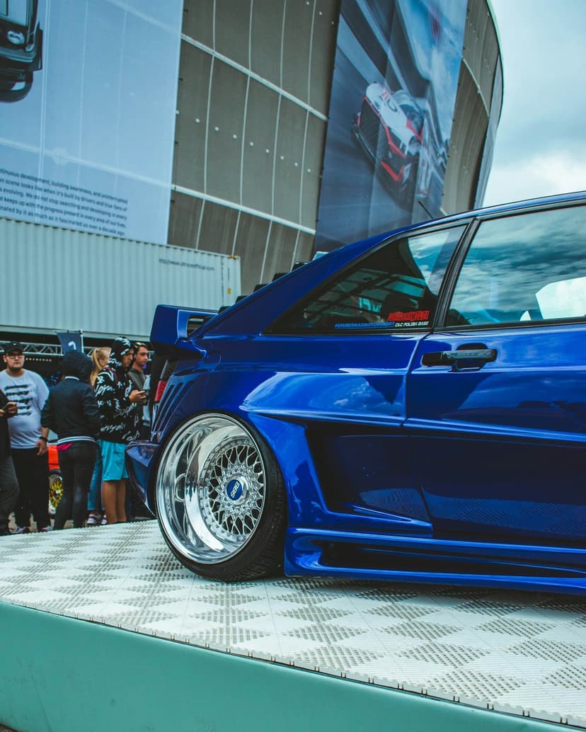 Blue Audi Sport Quattro with widebody fenders and polished mesh rims on a display platform at an outdoor show.