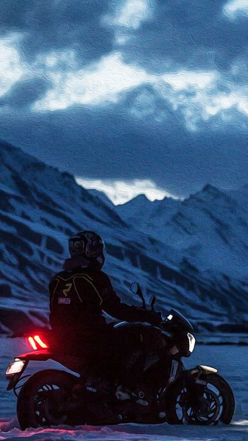 Motorcyclist on a dark bike faces jagged snow-covered mountains under a heavy layer of dark, swirling clouds.