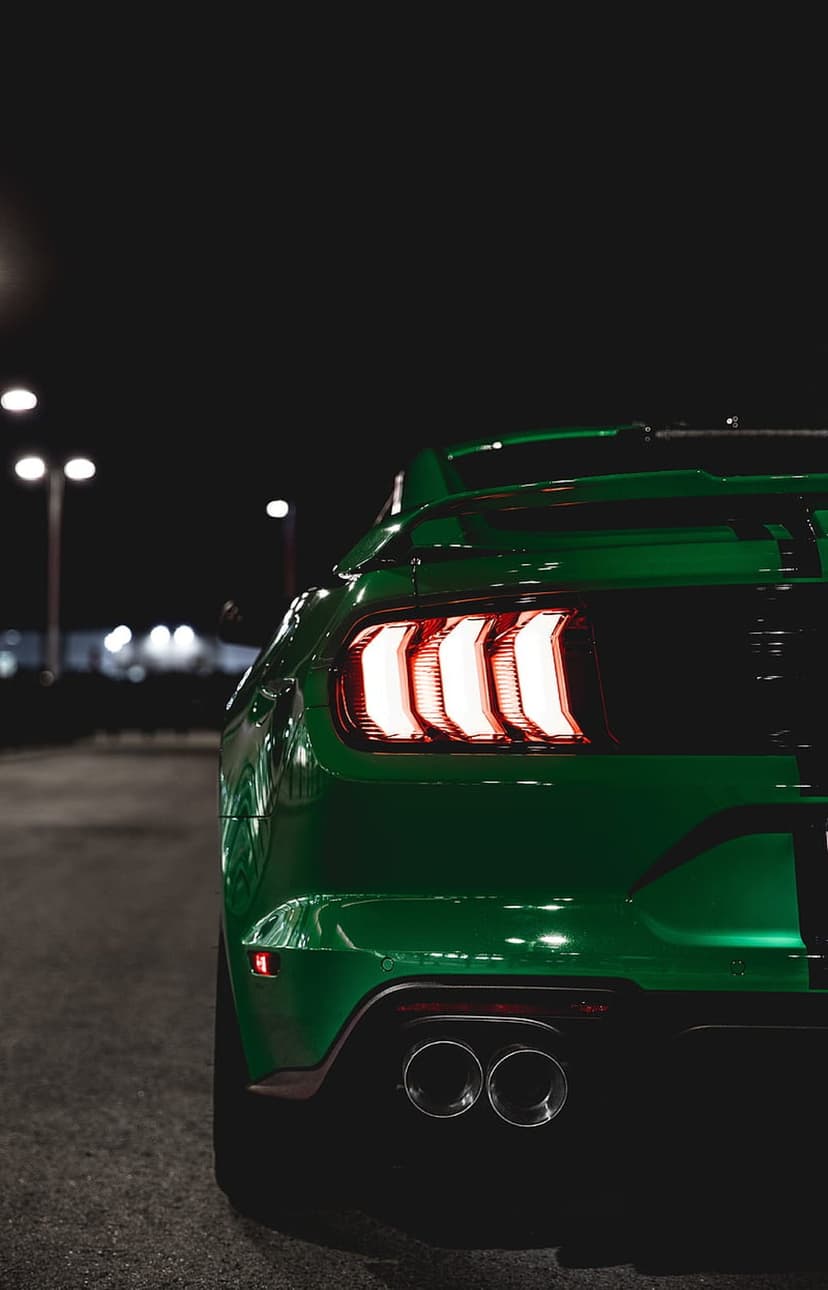 Green Ford Mustang tail lights glow red at night with dual exhaust pipes visible on dark asphalt pavement.