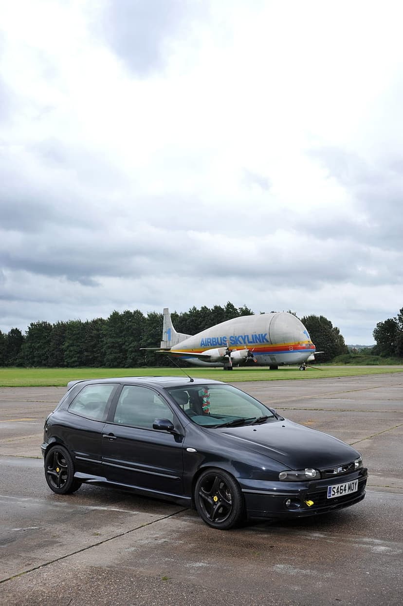 Black Fiat Bravo parked on an airfield with a large white Airbus Beluga cargo plane under a cloudy sky.