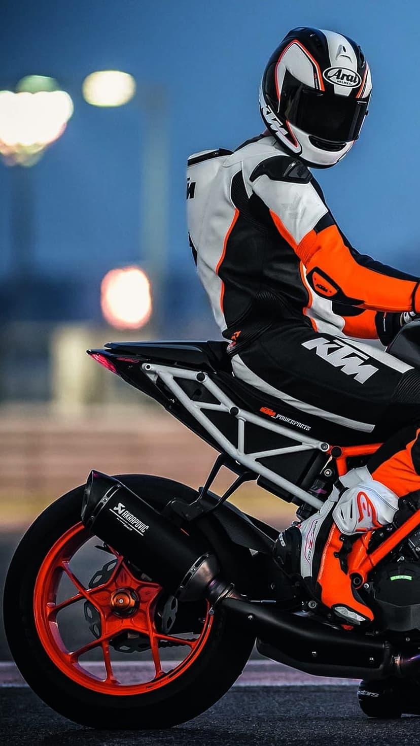 KTM motorcycle racer in an orange suit sits on a sportbike with an Akrapovic exhaust on a blurred race track.