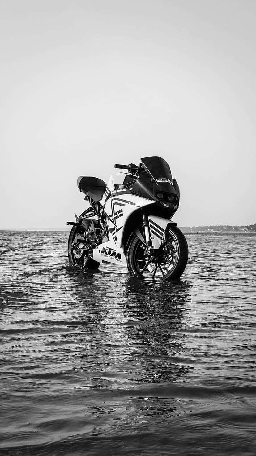 KTM motorcycle stands partially submerged in rippling coastal water under a bright, high-contrast monochrome sky.