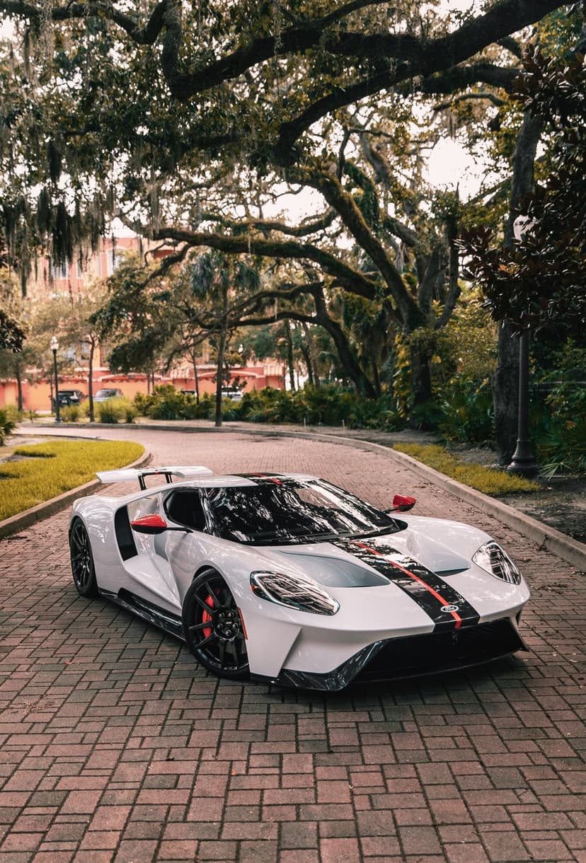 White Ford GT supercar with red racing stripes parked on a curved cobblestone path under mossy green trees.