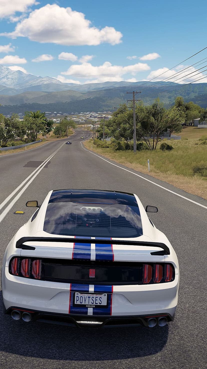White Ford Mustang with racing stripes drives on a winding road with mountains and a sunlit sky in the background.