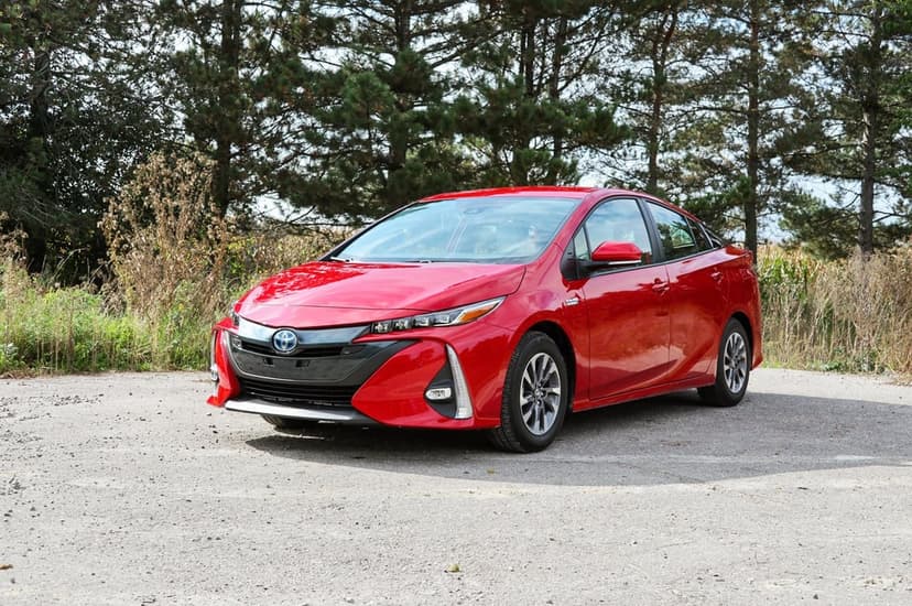 Red Toyota Prius Prime hybrid sedan parked on a gravel road with a background of dense green pine trees.