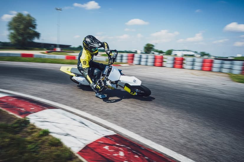 Motorcyclist on a Husqvarna supermoto leans into a racetrack curve with motion blur under a bright blue sky.