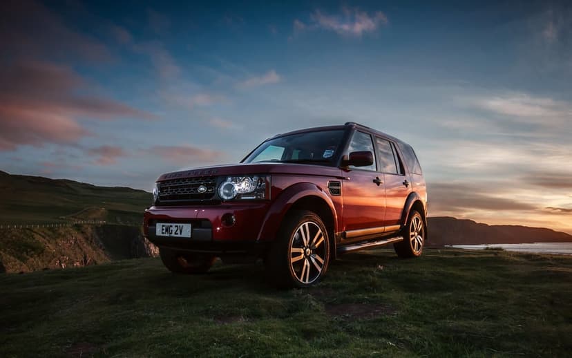 Red Land Rover SUV parked on a grassy cliff overlooking a rugged coast under a dramatic pink and blue sunset sky.