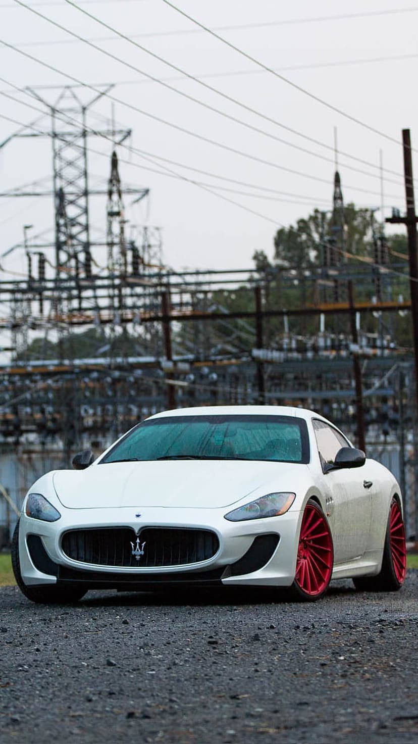 White Maserati GranTurismo with red wheels parked on gravel beneath industrial power towers and an overcast sky.