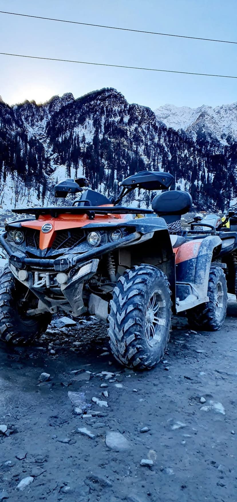 Orange and black ATV on a rocky ridge with snow-covered mountain peaks under a clear blue alpine winter sky.