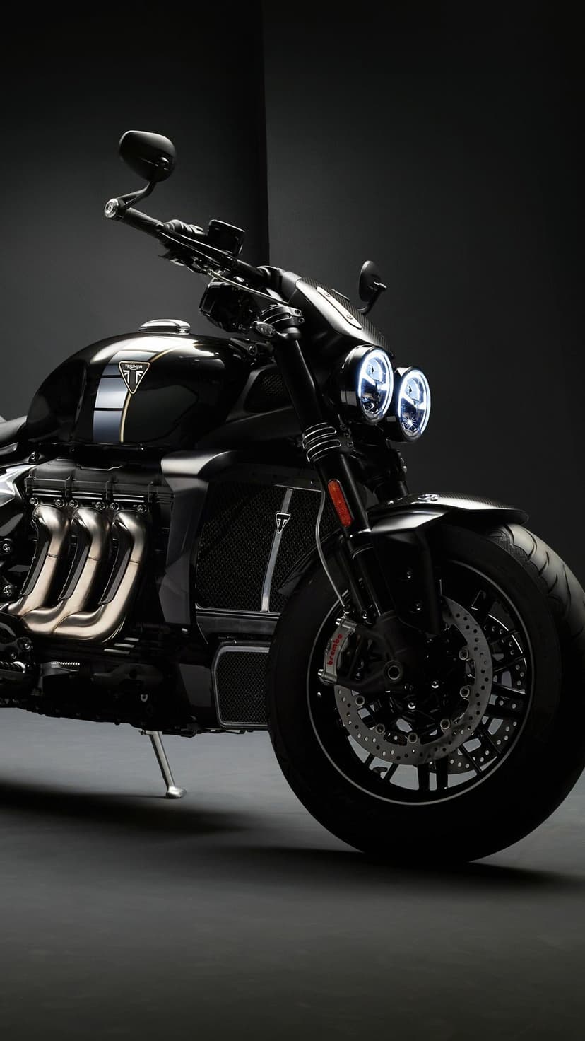 Black Triumph motorcycle with twin LED headlights and chrome exhaust pipes in a dark studio setting.