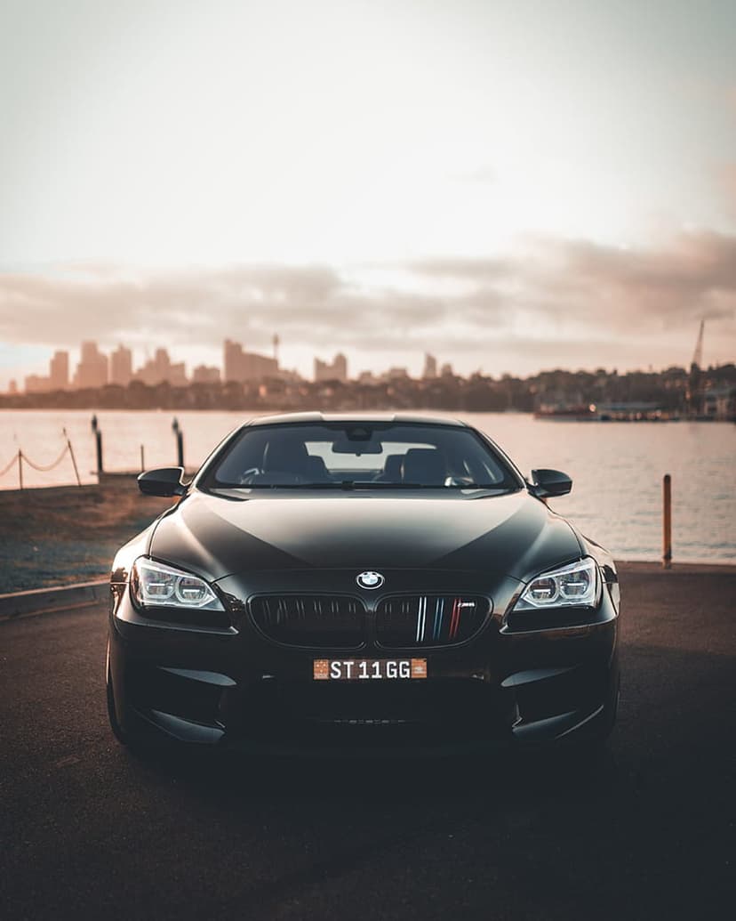 Black BMW M6 convertible parked on a coastal road during sunset with glowing headlights and a city background.