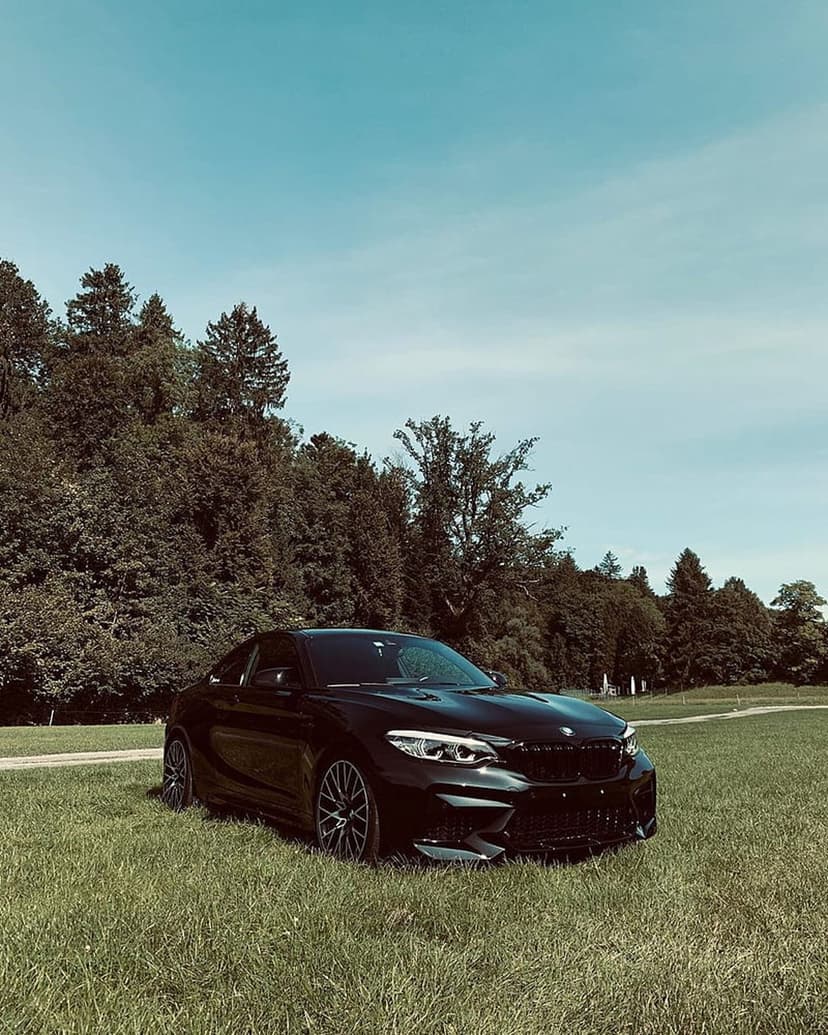 Black BMW M2 sports car parked on a green grass field with a backdrop of tall trees under a bright blue sky.