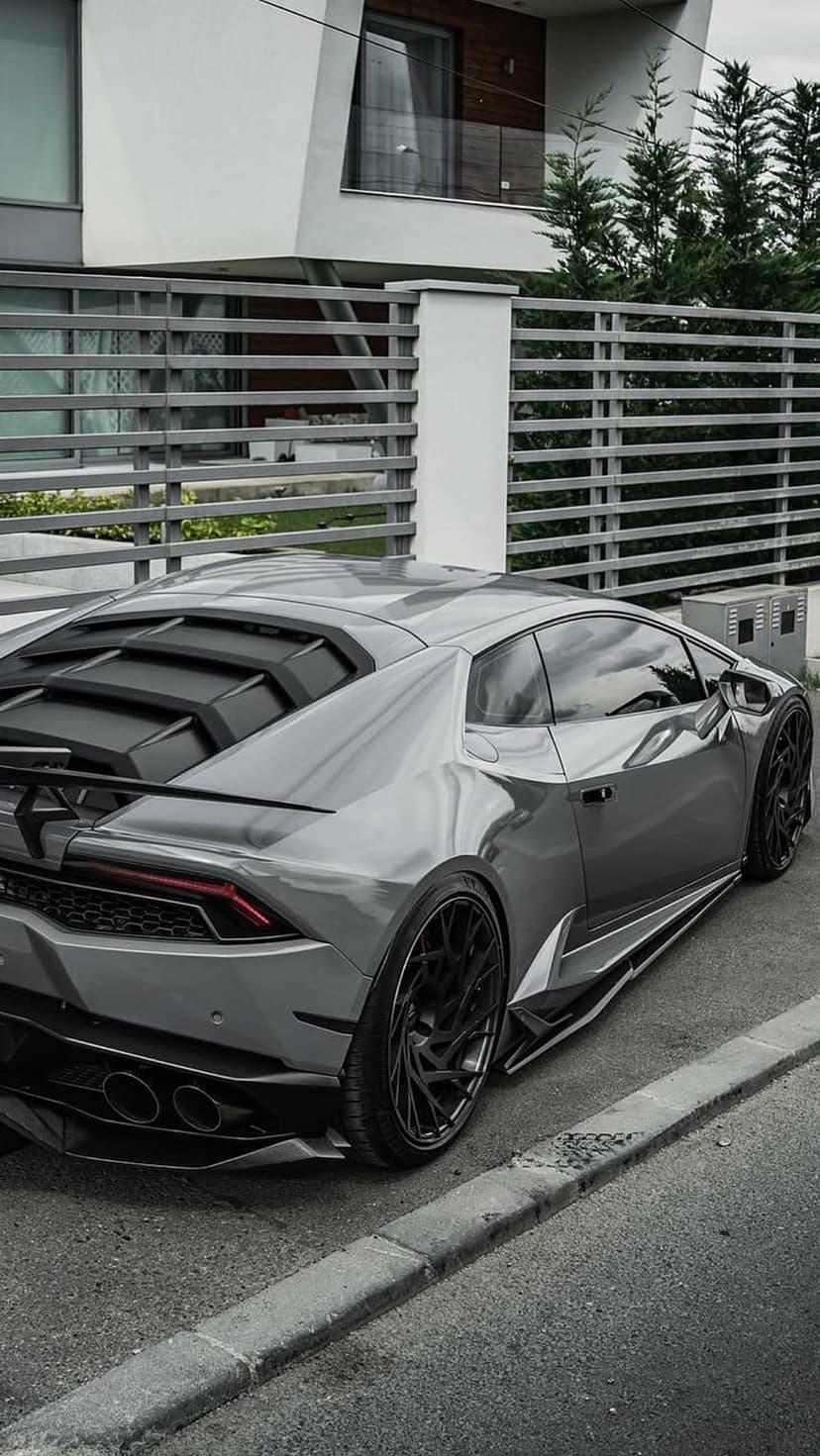 Grey Lamborghini sports car with black alloy wheels parked in the driveway of a sharp-angled modern luxury home.