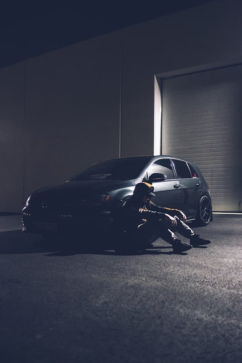 Man sitting beside a dark car in a dimly lit industrial garage with dramatic shadows and concrete walls.