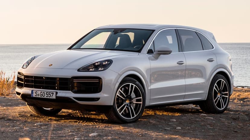 Silver Porsche Cayenne SUV parked on a sandy beach during sunset with calm ocean waves in the background.
