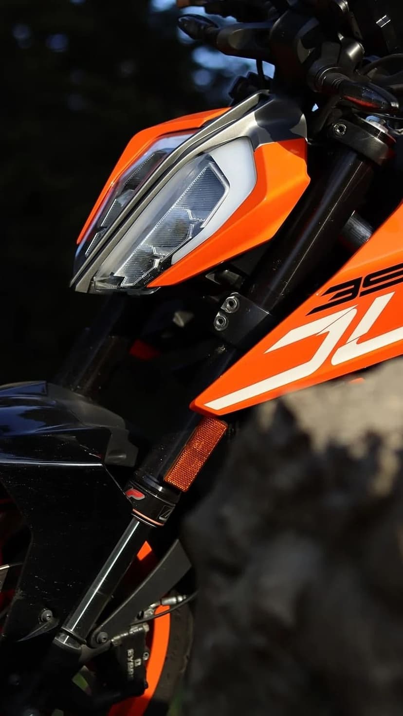 KTM 390 Duke motorcycle front with split LED headlight, orange fairings, and black forks against a rocky hill.