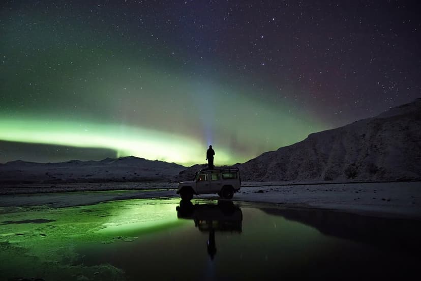 Person on a car beams a flashlight at green northern lights reflected in a calm lake surrounded by snow.