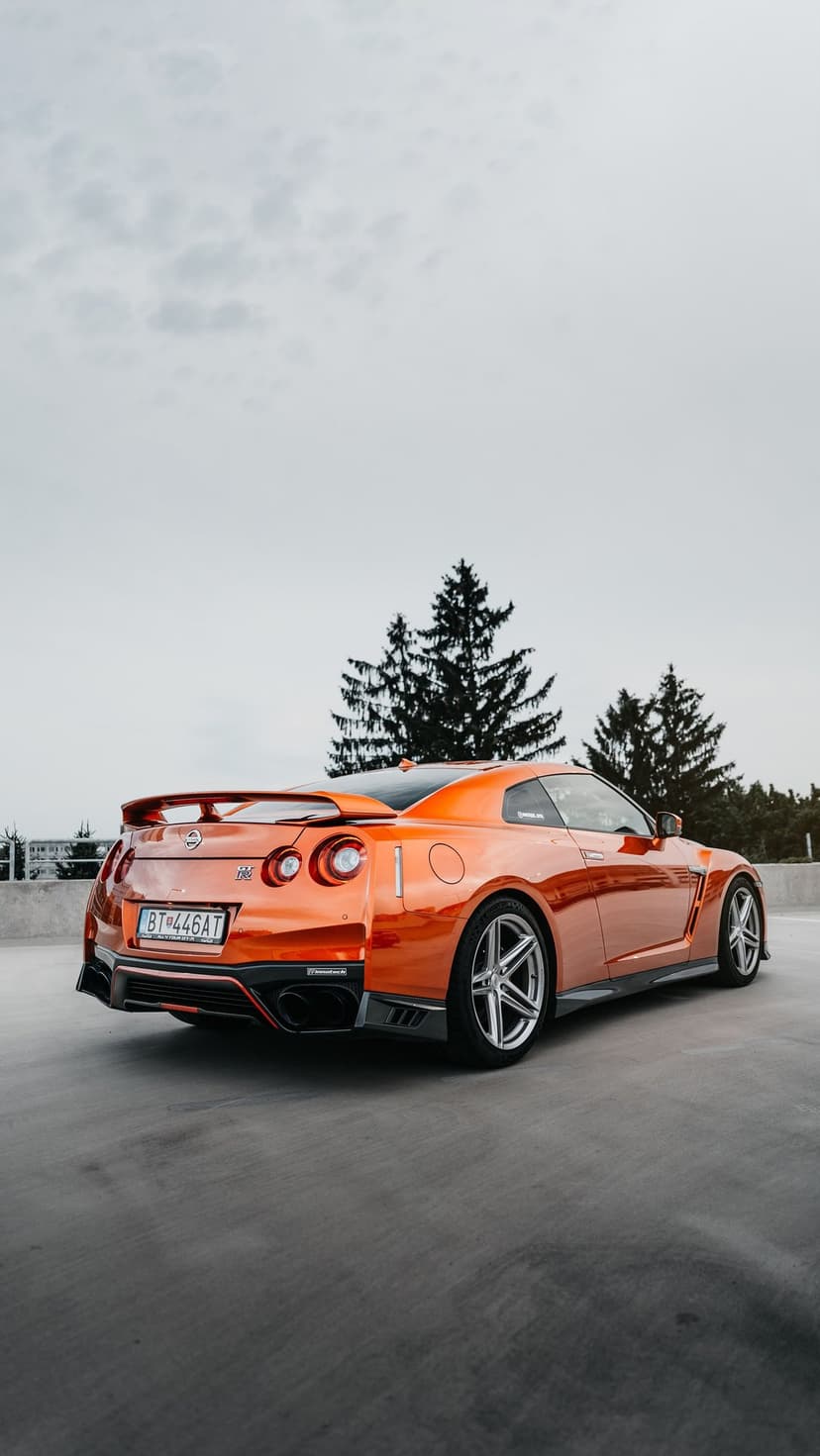 Nissan GT-R in metallic orange parked on concrete with rear taillights visible and a backdrop of tall pine trees.