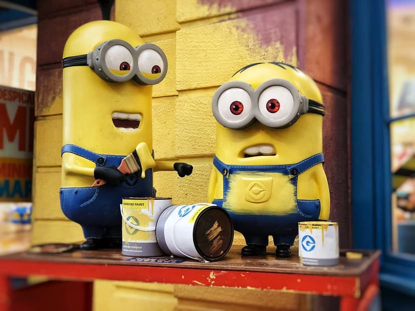 Two Minions in denim overalls handle yellow paint buckets and brushes amidst vibrant splatters and spills.