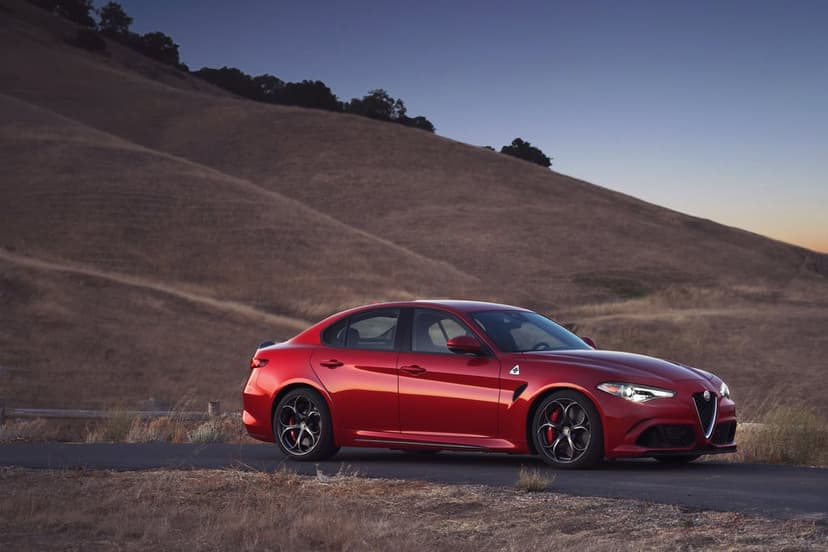 Red Alfa Romeo sports sedan parked on a paved road at twilight with dry rolling hills in the background.