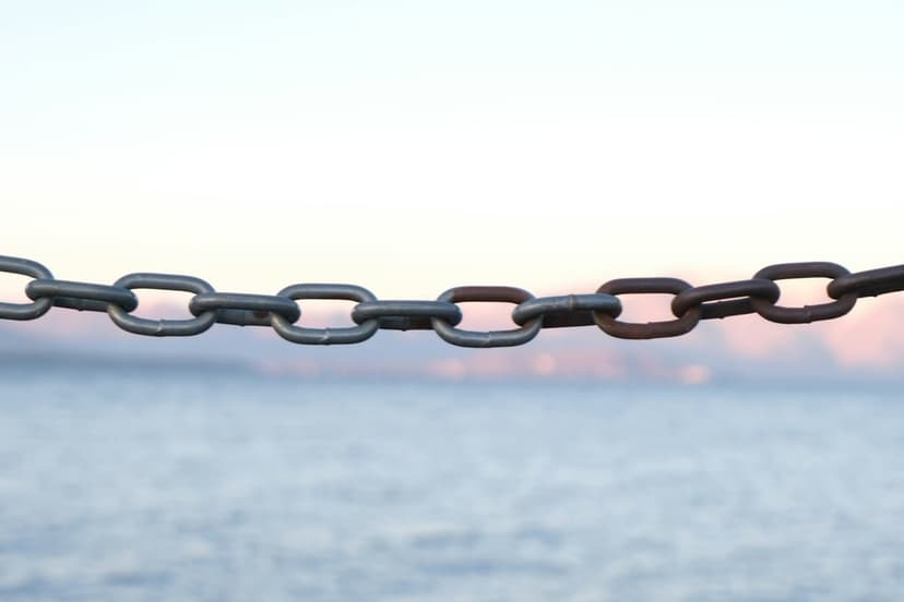 Oxidized metal chain links stretch across a calm blue sea under a soft pink and orange sunset horizon.