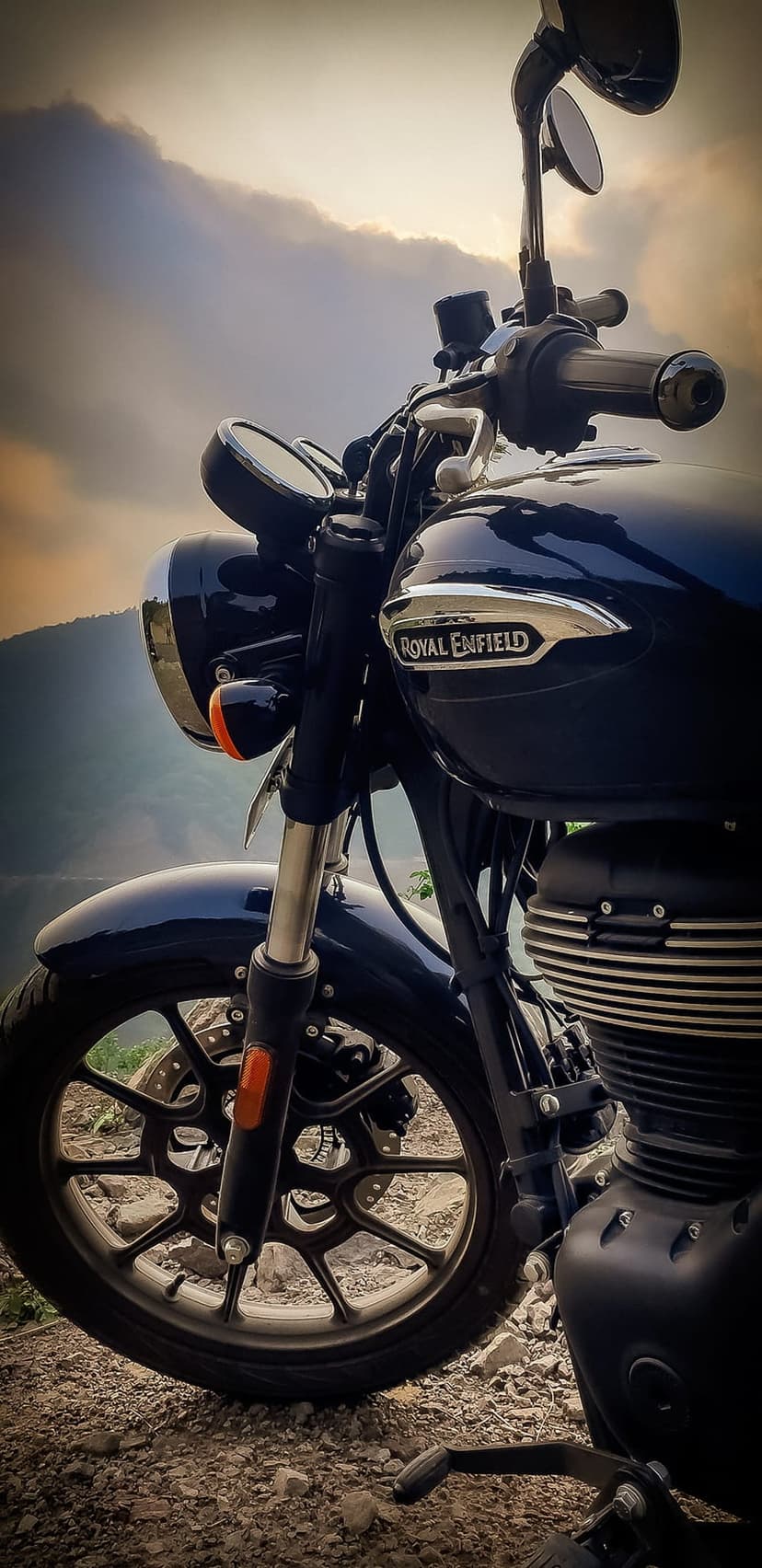 Navy blue Royal Enfield motorcycle on a rocky mountain road under a cloudy dusk sky with focused chrome engine parts.