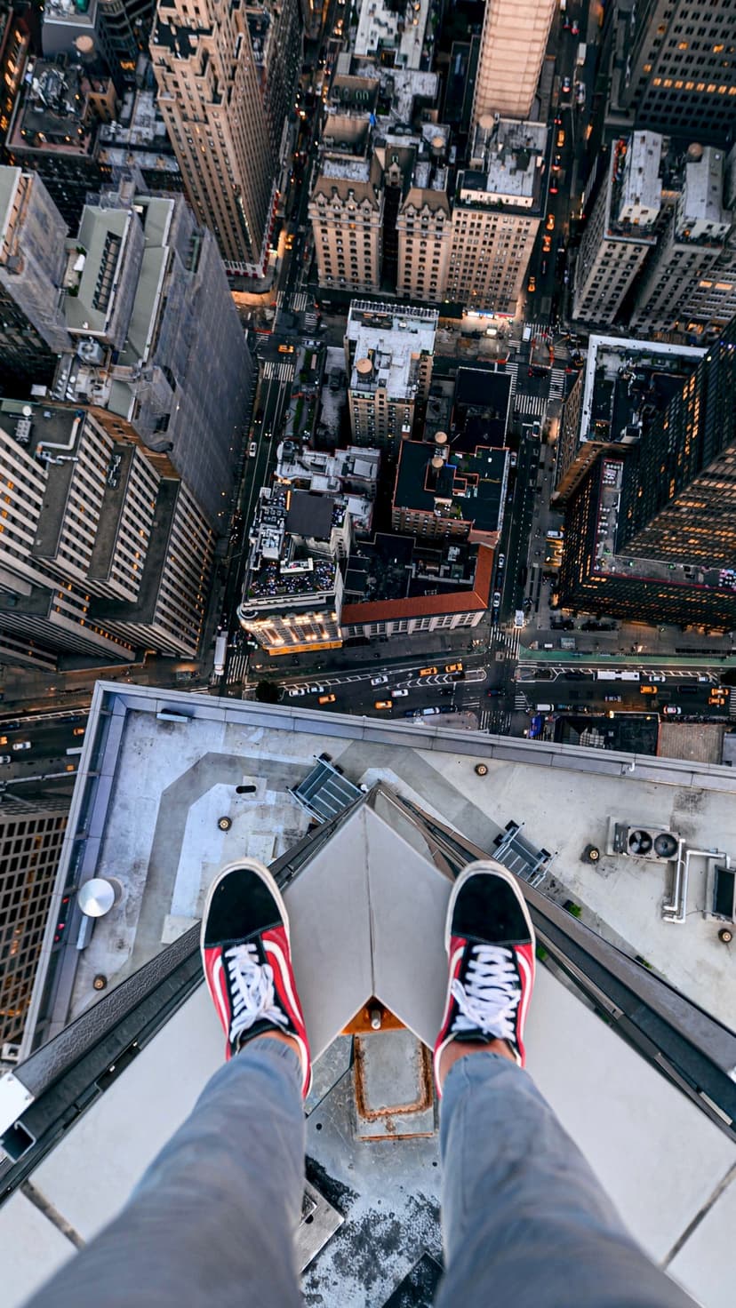 Sneakers on a narrow skyscraper ledge overlook a glowing night cityscape with countless street lights and buildings.
