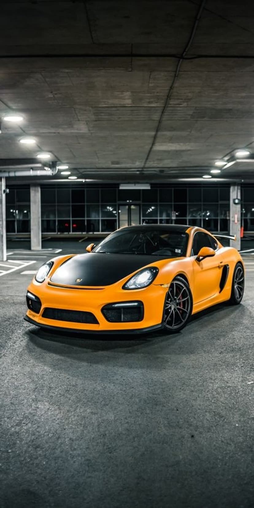 Orange Porsche Cayman GT4 with a matte black hood and glowing headlights parked in a dark concrete garage.