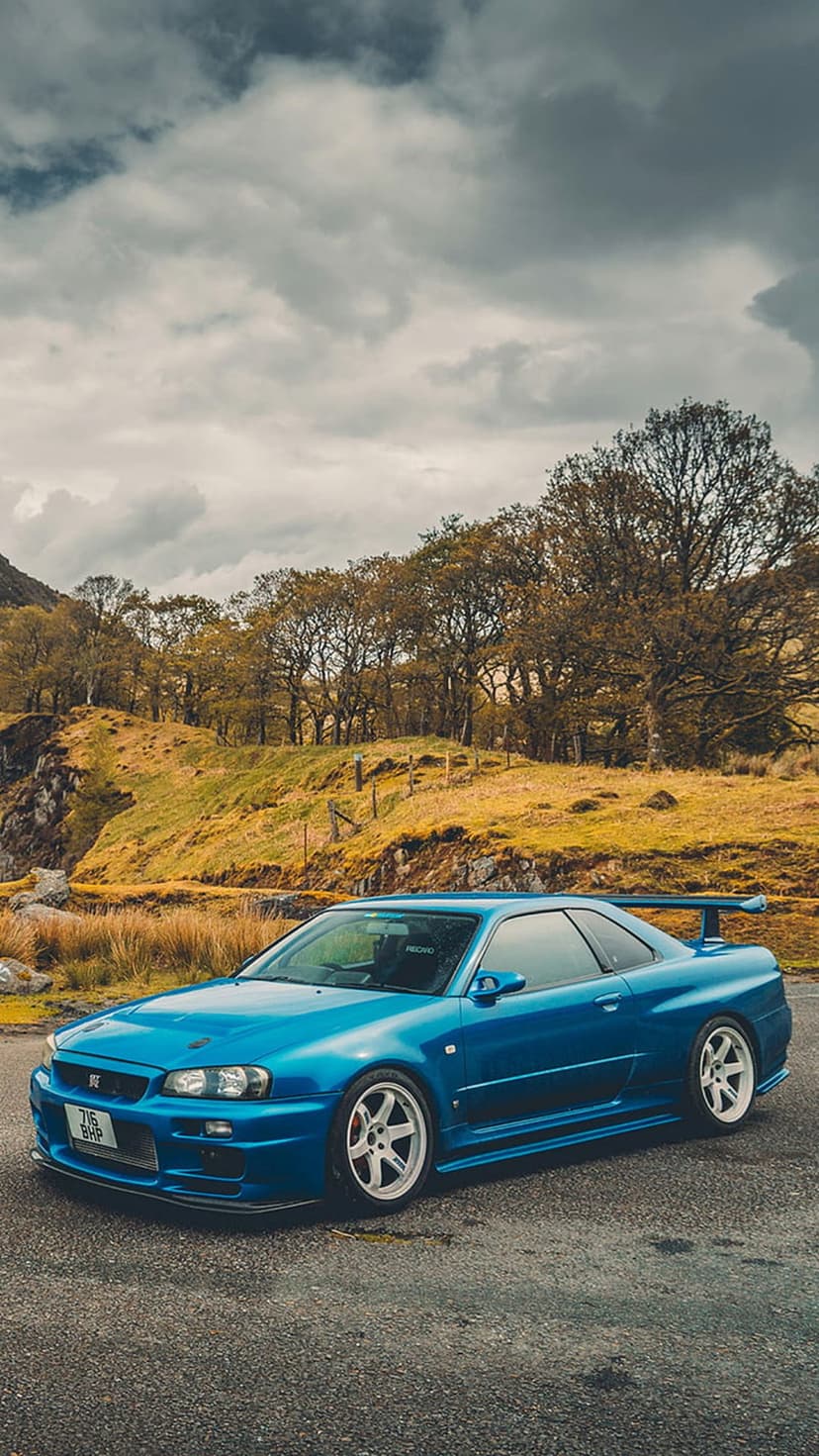 Blue Nissan Skyline R34 GT-R with white wheels parked on a paved road under a cloudy sky near autumnal hills.