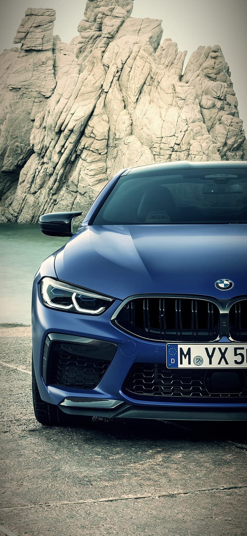 BMW M8 sports coupe in metallic blue parked on gray coastal rocks with a calm ocean horizon in the background.