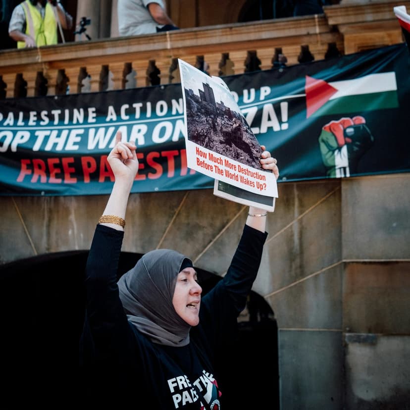 Woman in a black hijab holds a protest sign showing urban destruction at a Free Palestine rally outdoors.