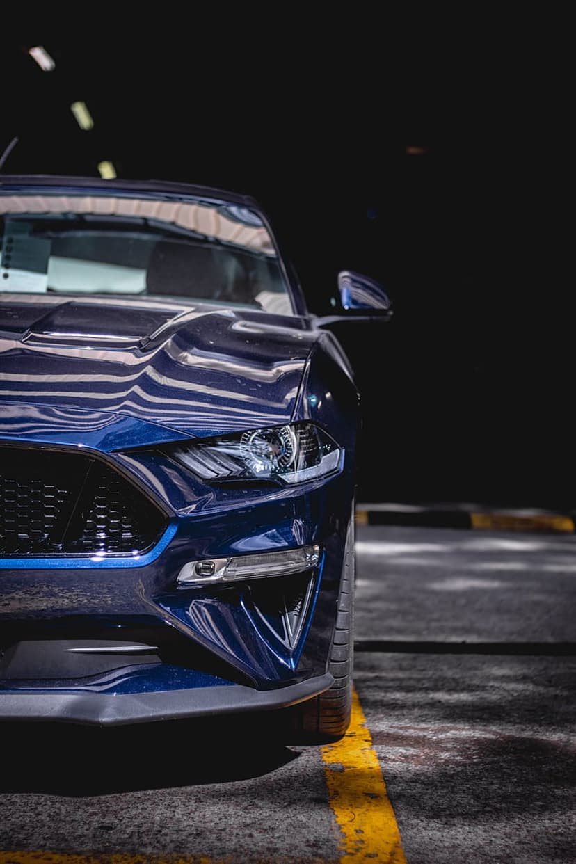 Blue Ford Mustang headlight and grille close-up with glossy reflections and blurred garage background.