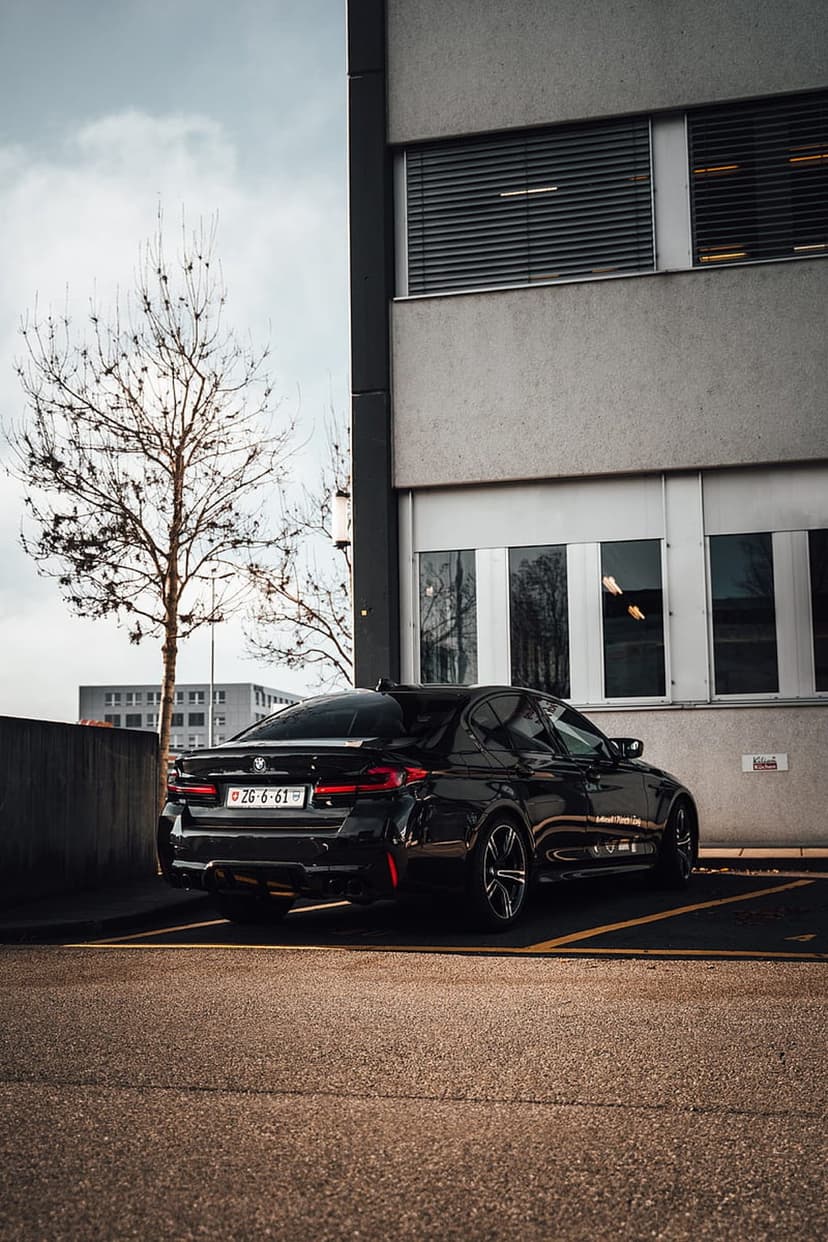 Black BMW M5 sedan parked in an urban lot featuring yellow lines and a modern grey glass building background.