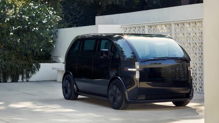 Black futuristic electric van parked by a white lattice wall and green bushes under bright sunlight.