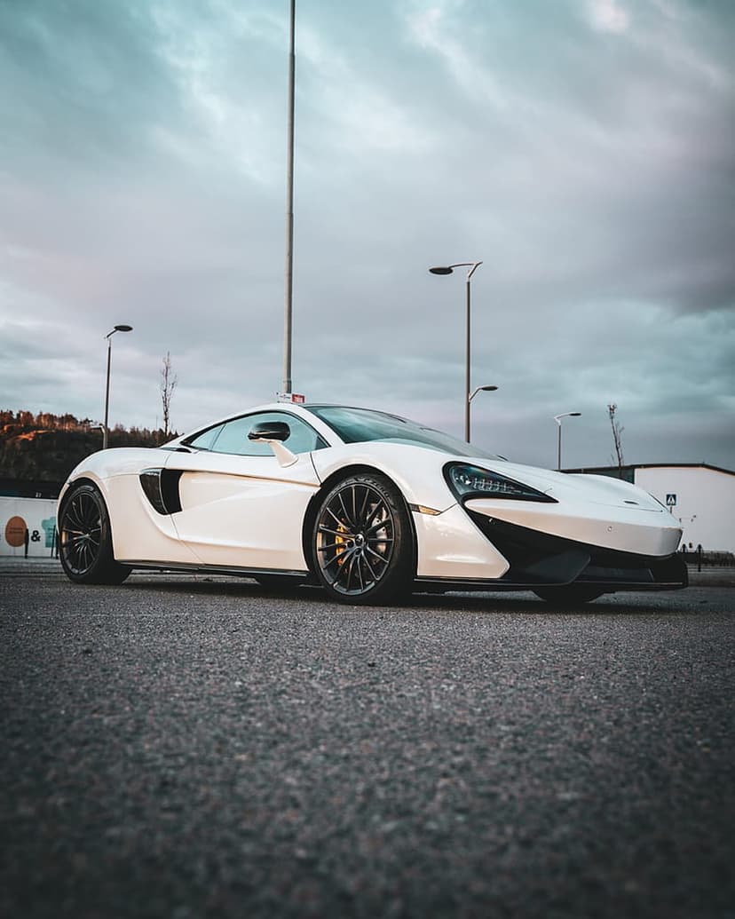 White McLaren 650S supercar parked in an asphalt lot under a dark cloudy sky during dusk at a low angle.