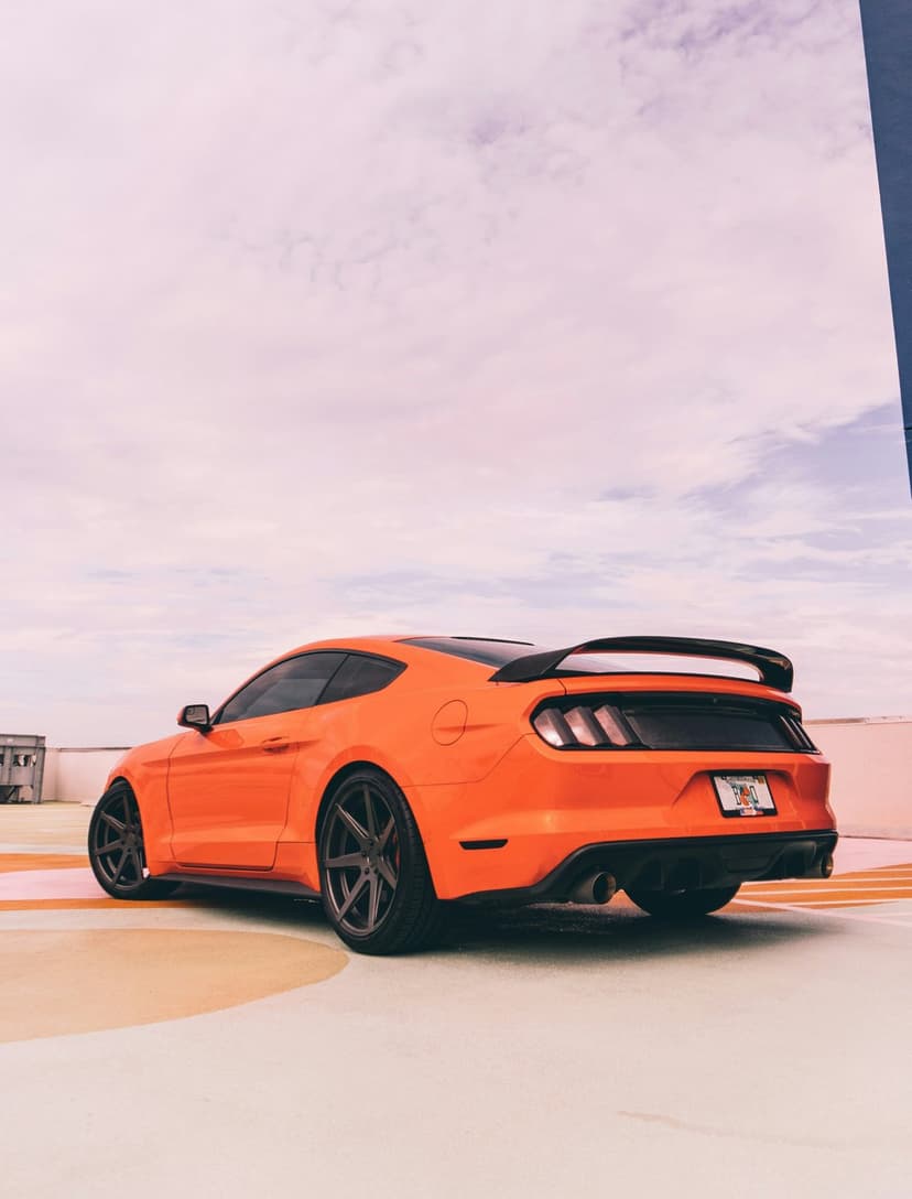 Orange Ford Mustang GT with a rear spoiler and dark wheels parked on a rooftop under a pink and white sky.