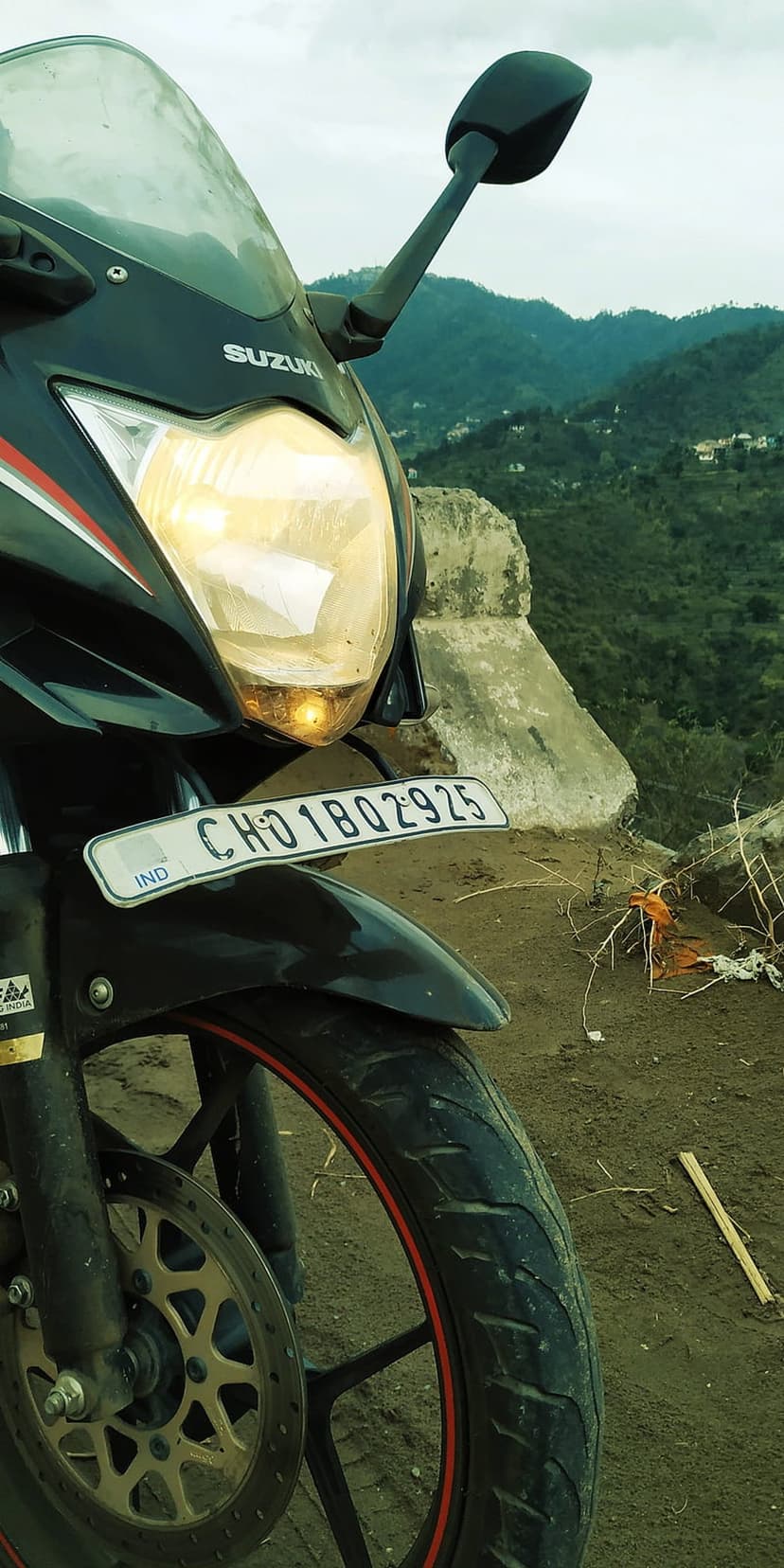Black Suzuki motorcycle headlight and Indian license plate against a backdrop of lush green mountain slopes.