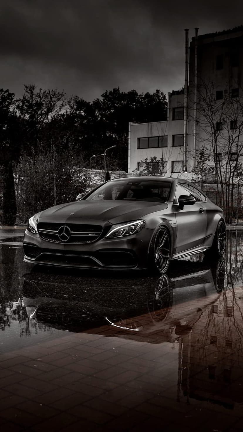 Matte black Mercedes-Benz coupe parked on wet asphalt with its reflection visible in a puddle under a cloudy sky.