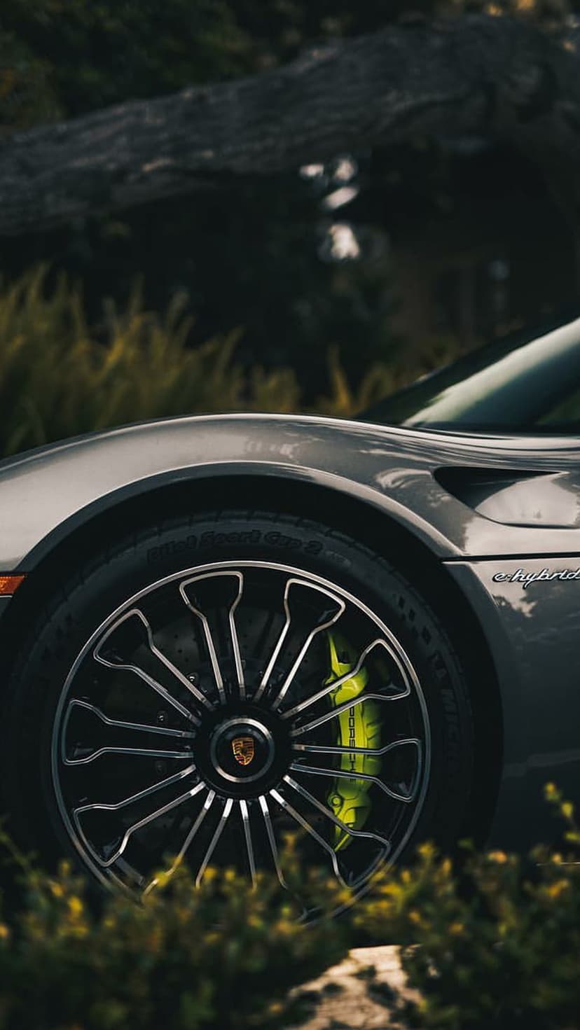 Porsche multi-spoke alloy wheel features bright green brake calipers and a hybrid badge against blurred trees.